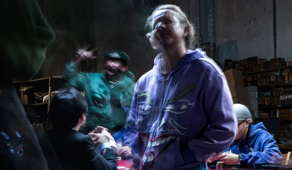 Laughing figure in the purple Joker face hoodie at a smoky poker game