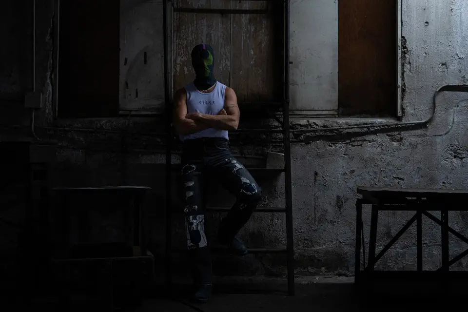 Bane mask and white tank leaning on a ladder in a derelict room