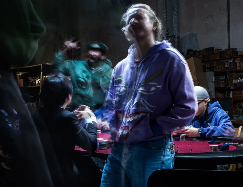 Joker face hoodie standing over a poker game in a warehouse