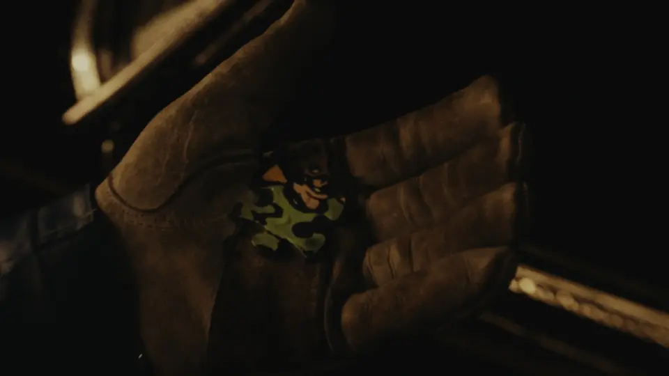 Film still: gloved hand holding a green Riddler puzzle piece